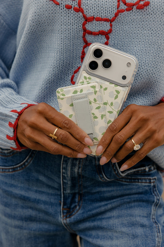Floral magnetic wallet with gray strap closure held in hands beside a phone