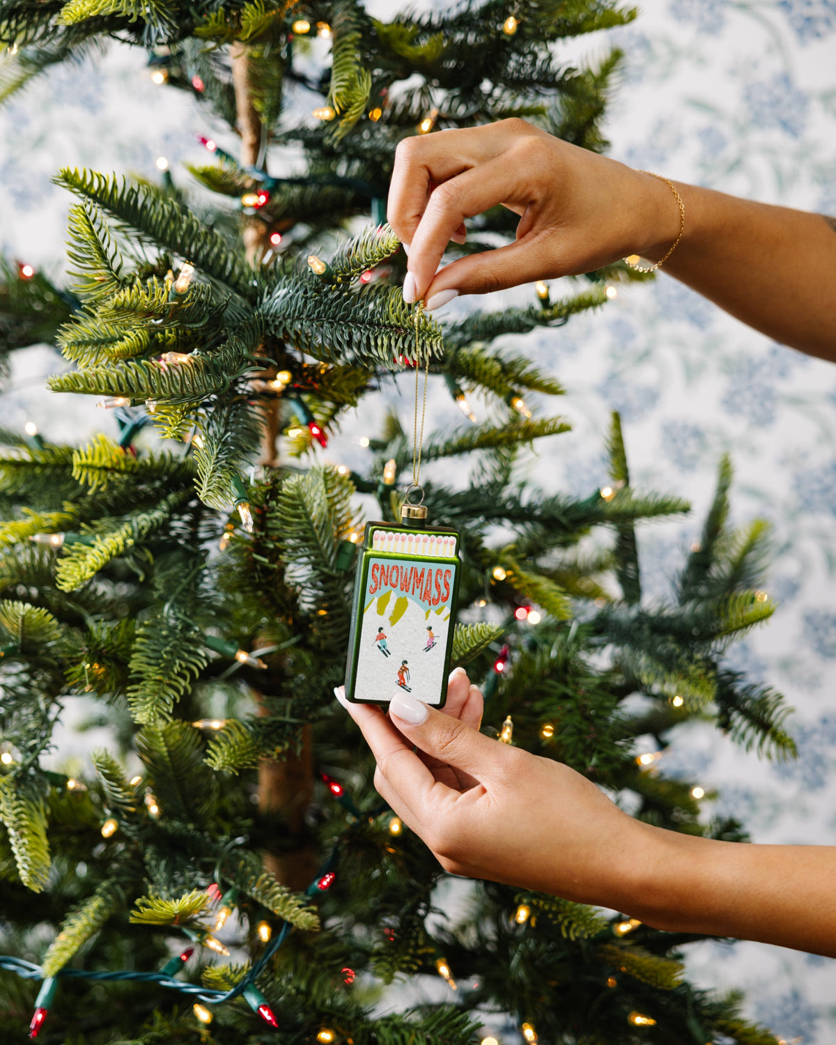 Snowmass matchbook ornament on a gold cord, being placed on a Christmas tree