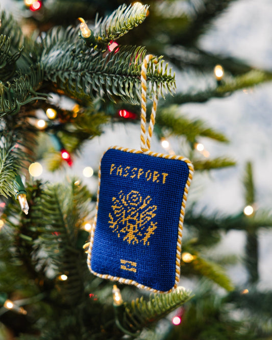 Blue needlepoint passport ornament hanging on a Christmas tree with gold stitched design.