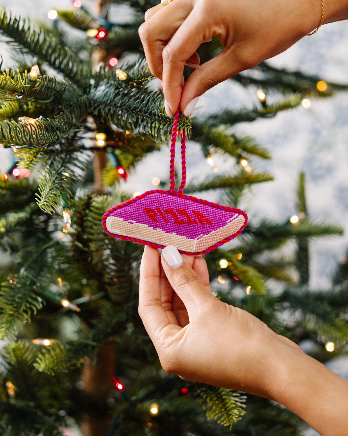 Needlepoint pizza slice ornament on pine branch as hands adjust the twine hanger