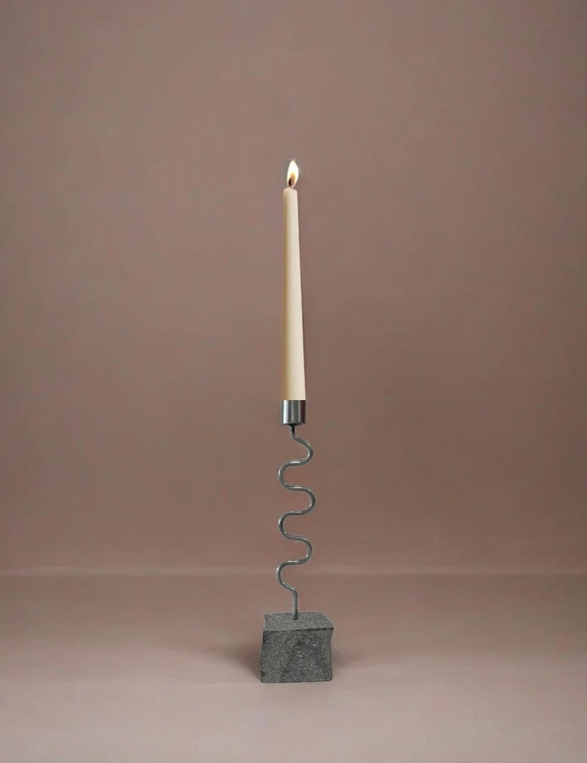 Candle holder with burning white candle on a wavy metal stand and dark gray base block