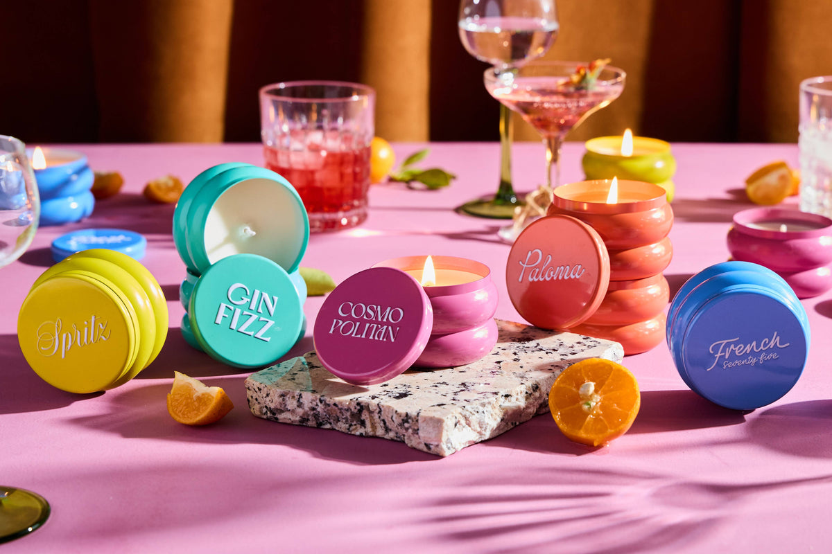 Rewined Cosmopolitan candle in an open pink jar among colorful candles; matching lid rests nearby.