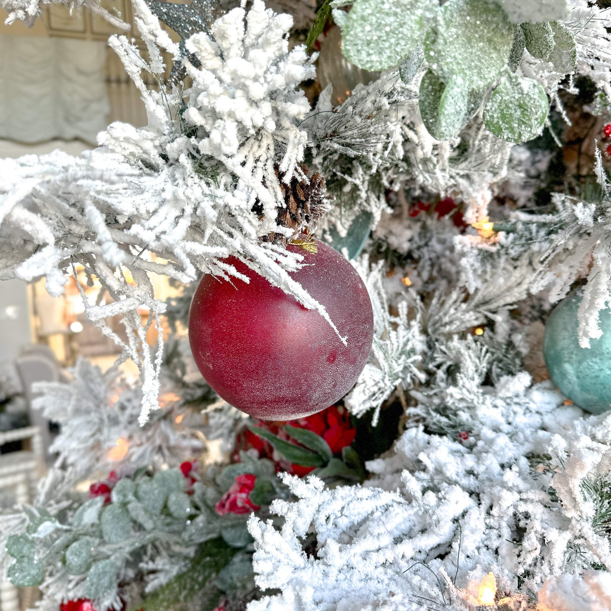 Red frosted glass ornament ball with hanging loop on a frosted Christmas tree.
