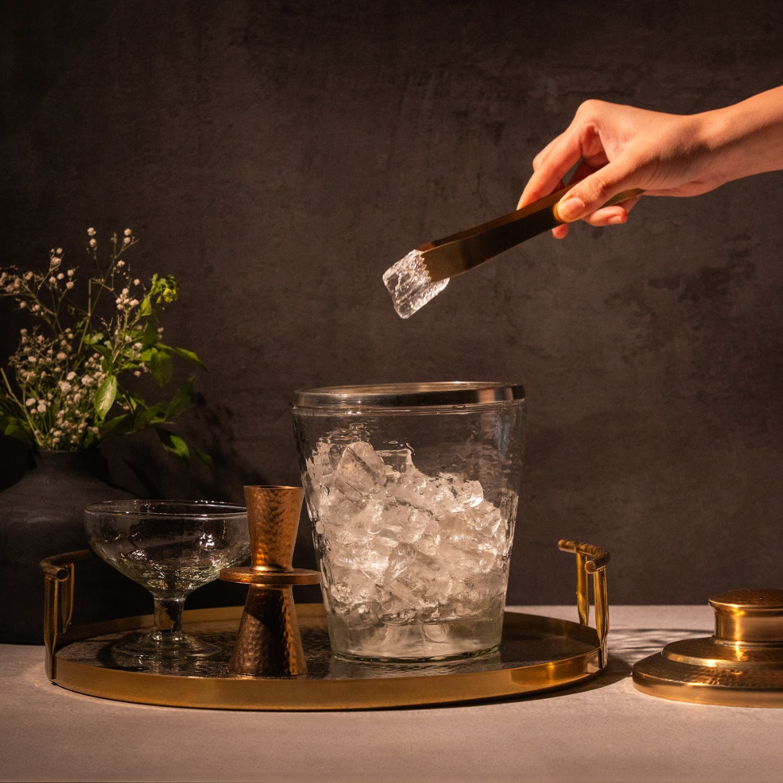 Hand holding gold tongs above a clear pebbled ice bucket on a gold tray