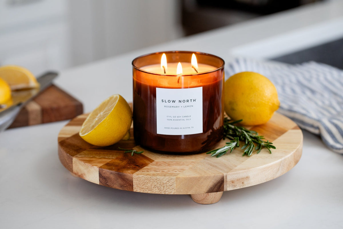 Amber jar three-wick candle with lit wicks, rosemary, and lemon on round wooden tray