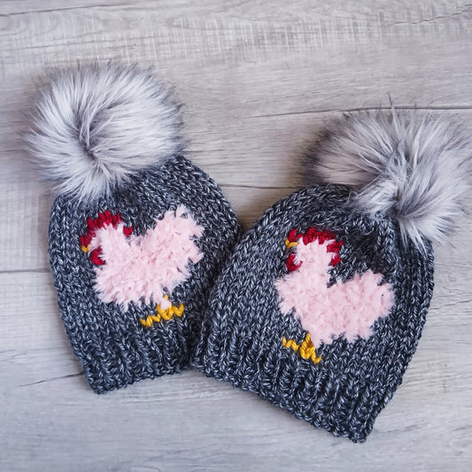Pair of slate knit beanies with light pink chicken patches and gray faux fur pom-poms
