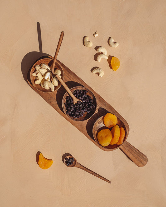 Wooden serving board with three round condiment bowls and two spoon utensils