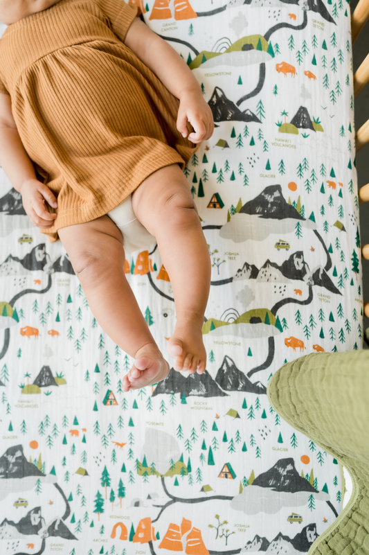 Crib sheet with white background and colorful national park illustrations; baby on a textured blanket.