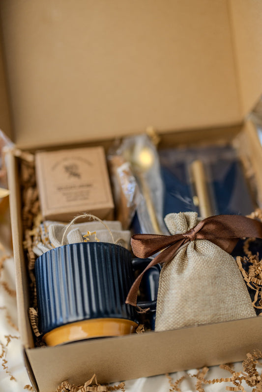 Gift box open view with navy ribbed mug, gold pen, and beige drawstring pouch