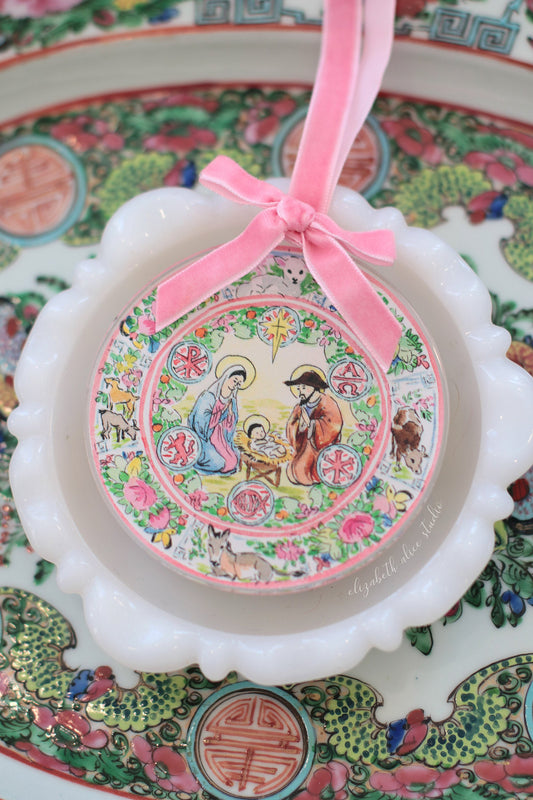 Nativity ornament with pink velvet bow, shown against a scalloped white edge