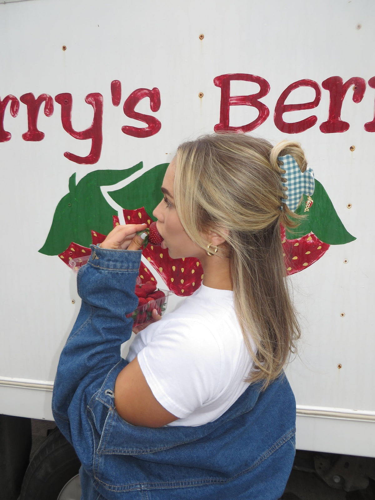 Blue gingham claw clip with red strawberry charm on blonde hair near wall sign.