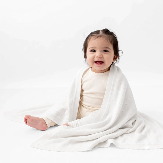 White pointelle knit baby blanket draped over baby, textured fabric with scalloped edge