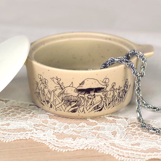 Beige ceramic trinket box with forest engraving on side; silver chain on fabric