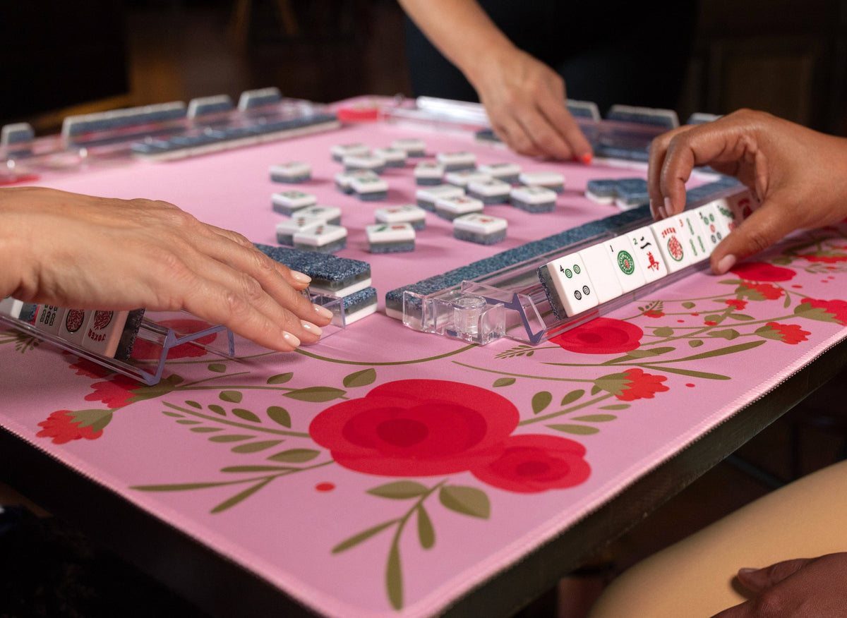 Pink floral mahjong mat on a table, tiles and hands arranging pieces