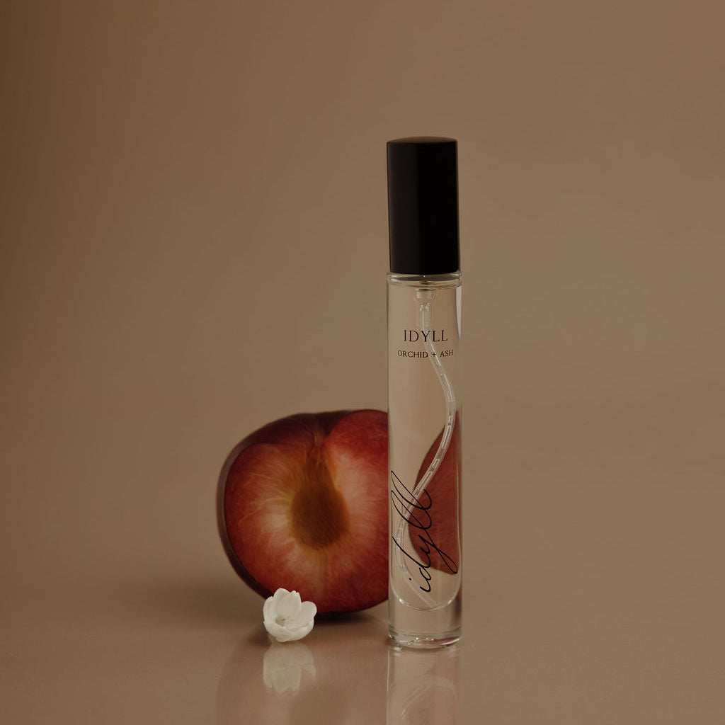IDYLL Orchid + Ash travel spray bottle with black cap beside red fruit and white flower
