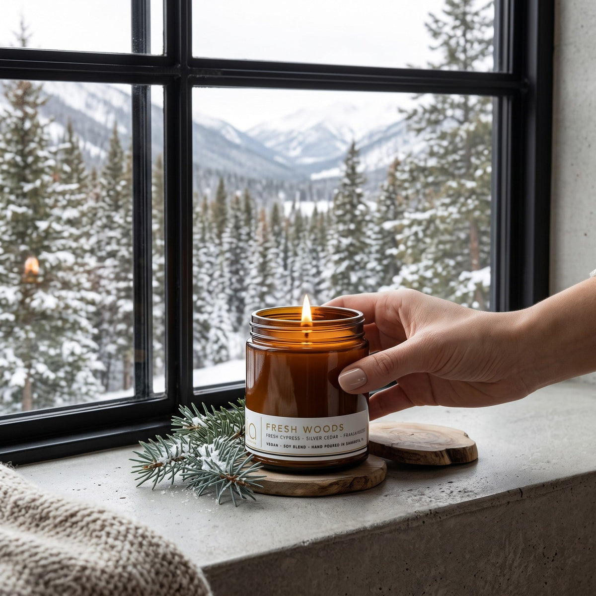 Hand holding lit Fresh Woods soy candle jar on a stone ledge with evergreen sprigs