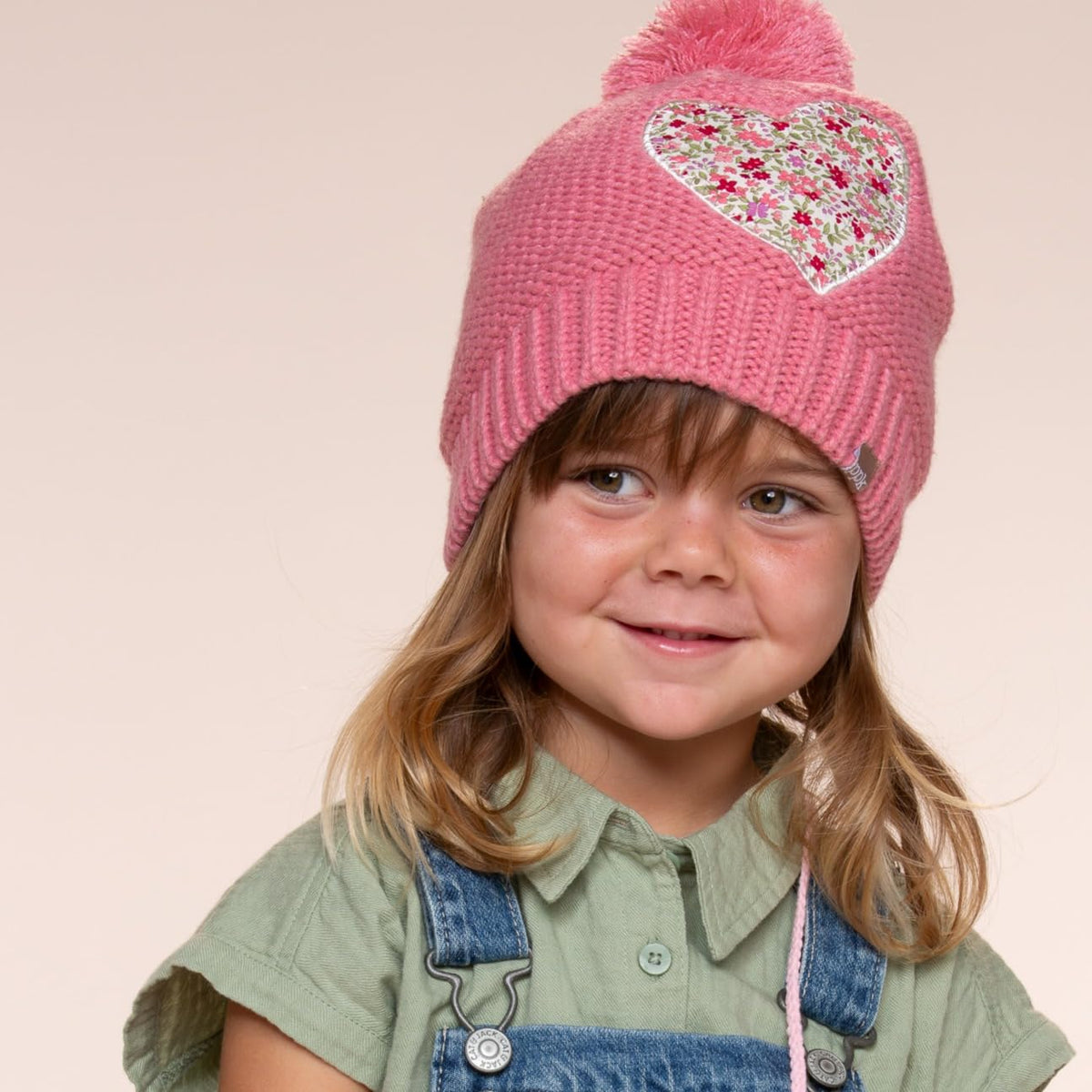 Child wearing a coral pink knit beanie with pompom and floral heart applique