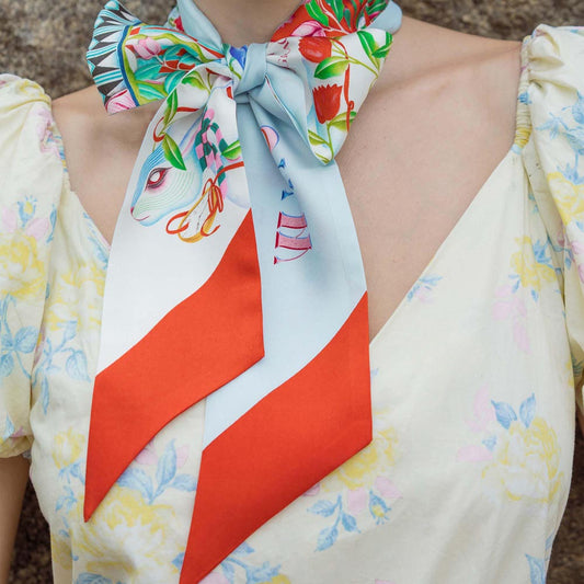 Silk twilly scarf worn at neckline, light blue with bird print and orange-red stripe