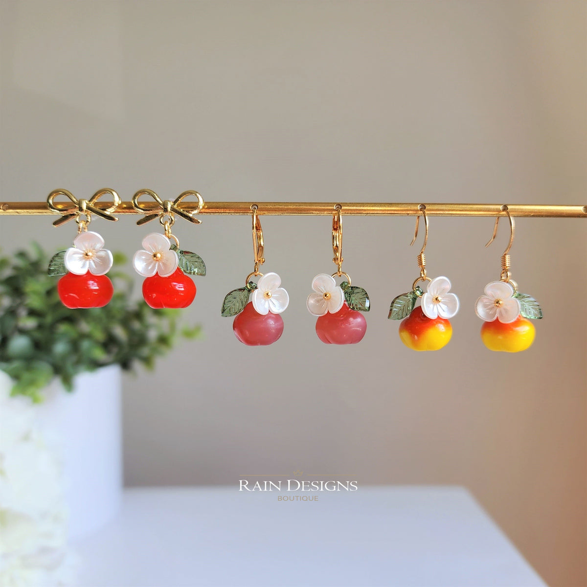 Gold apple-shaped dangle earrings on bar: red and yellow fruit charms with white flowers