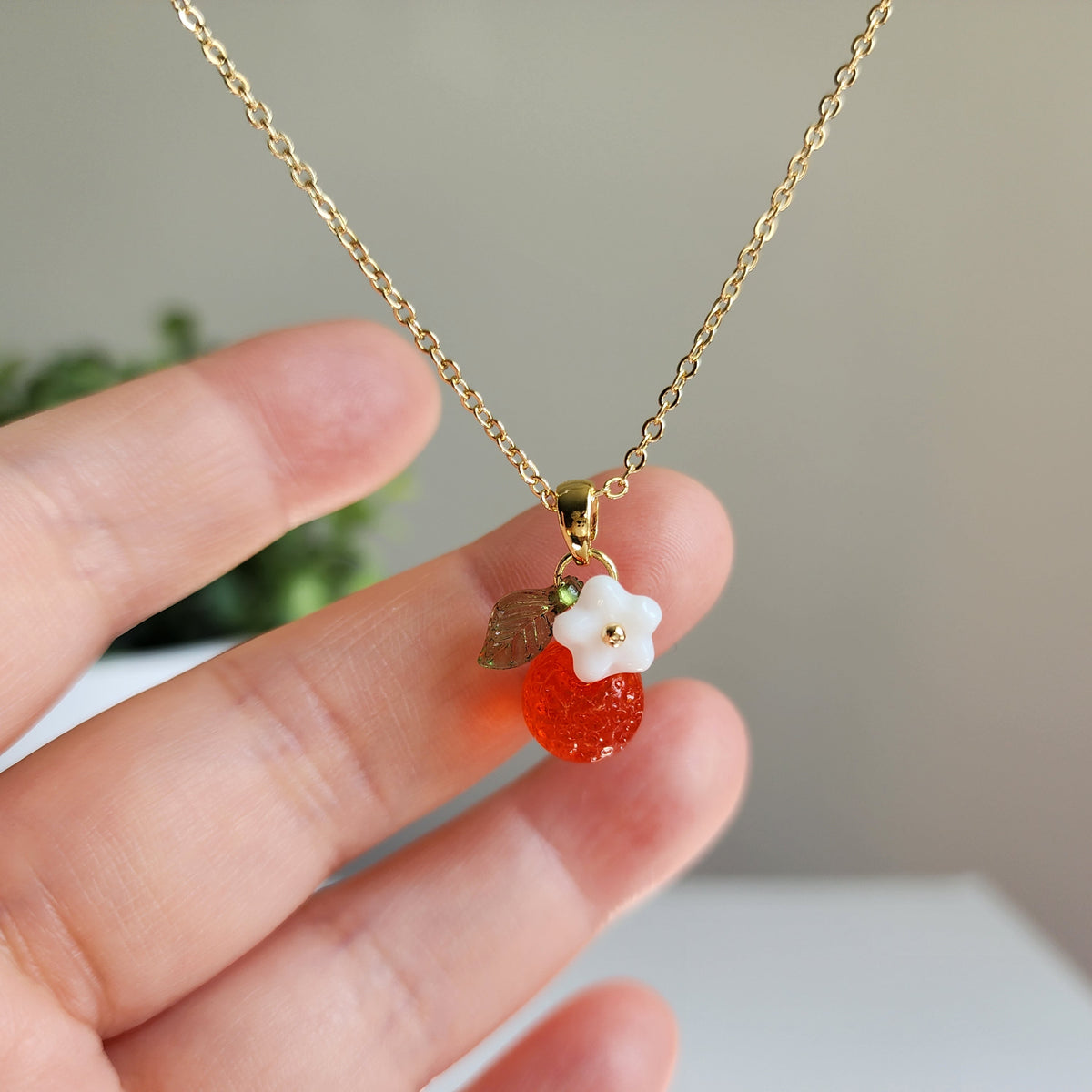 Gold-tone orange fruit charm necklace held in hand, with white flower and green leaf