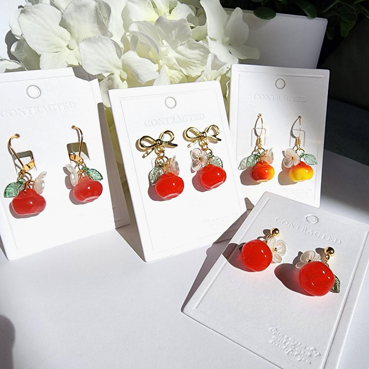 Gold apple earrings set on cards: red fruit charms with white flowers and green leaves