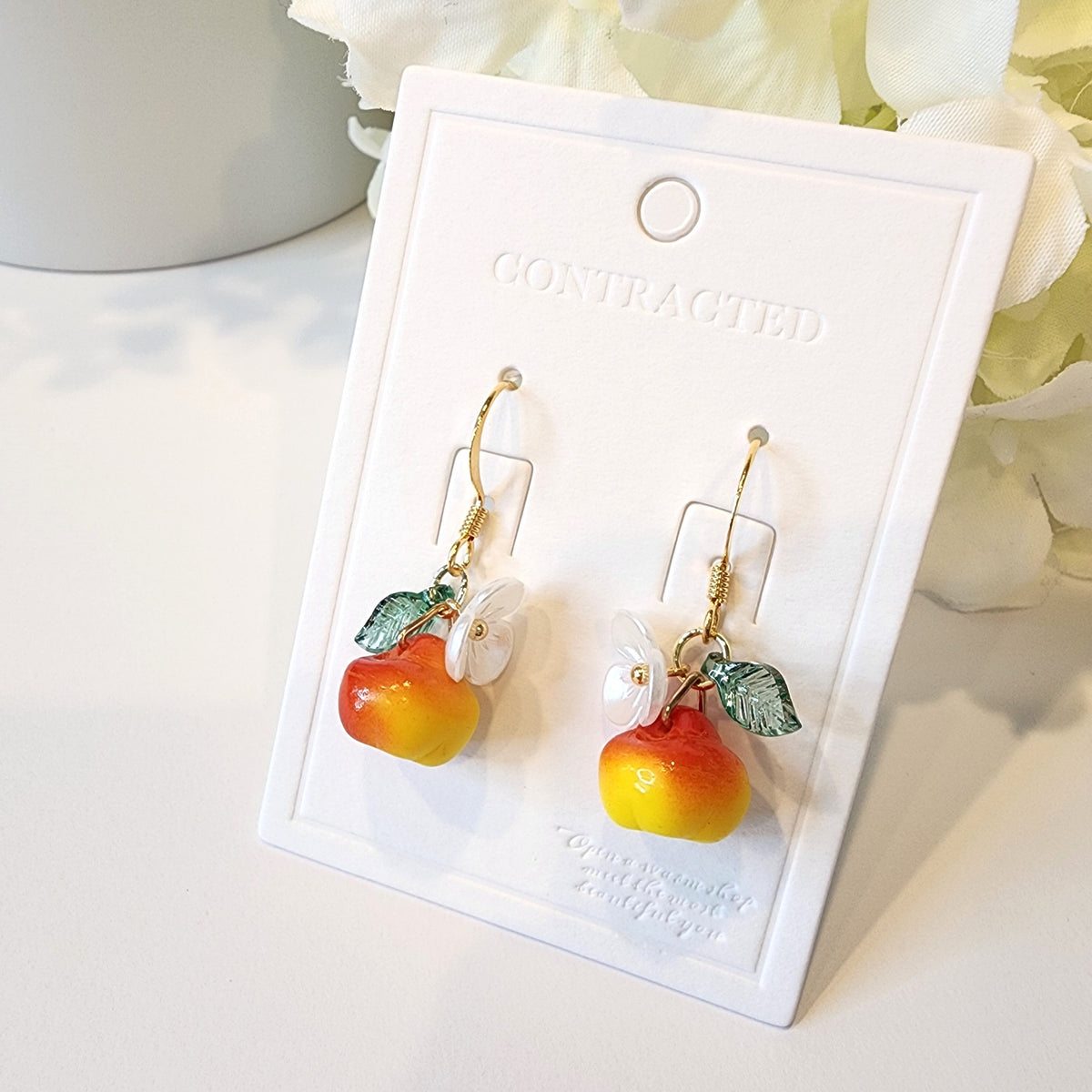 Gold hook apple earrings on card: red-yellow fruit charms with white flowers and green leaves