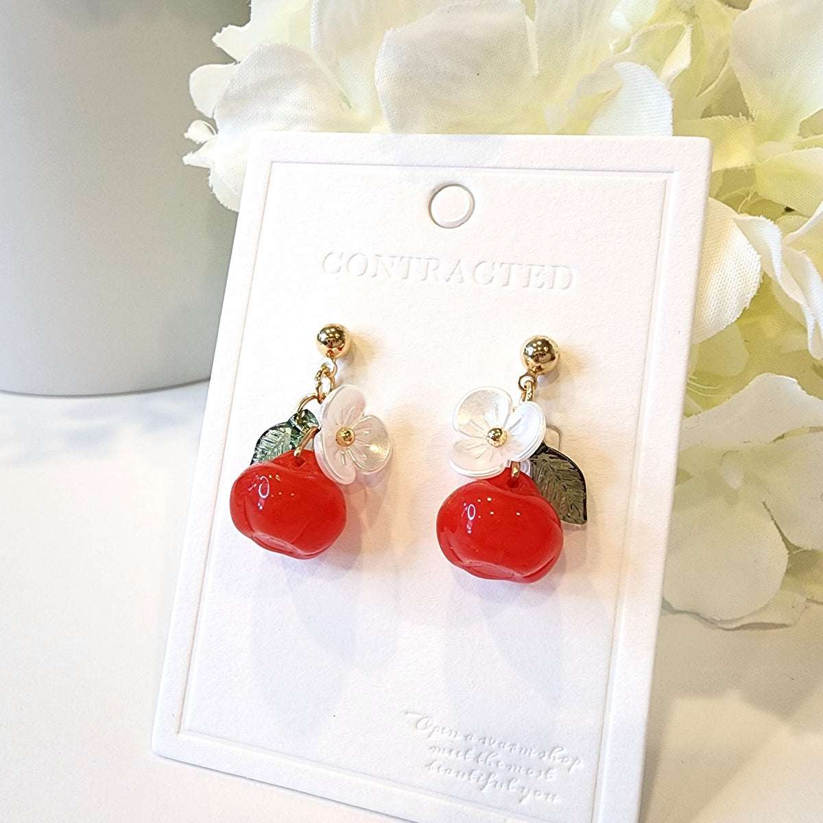 Gold hook apple earrings on card: red fruit charms with white flowers and green leaves