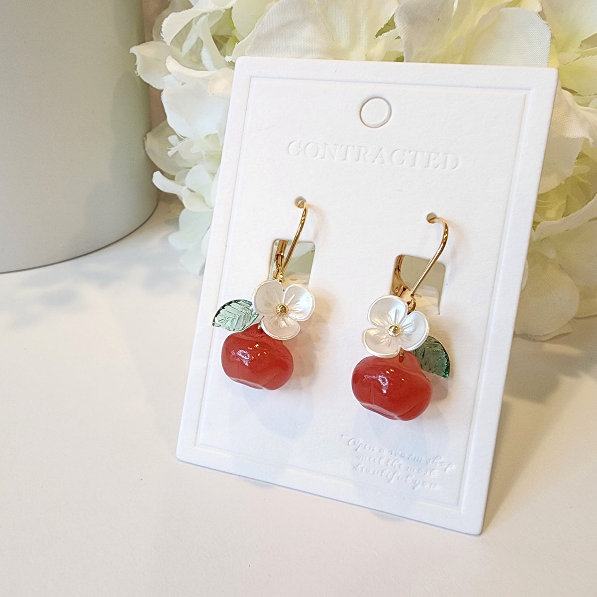 Gold hook apple earrings on card: red fruit charms with white flowers and green leaves