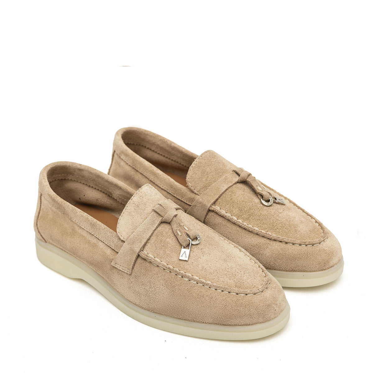 Pair of beige suede loafers with stitched aprons, strap detail, metal charms, and cream soles