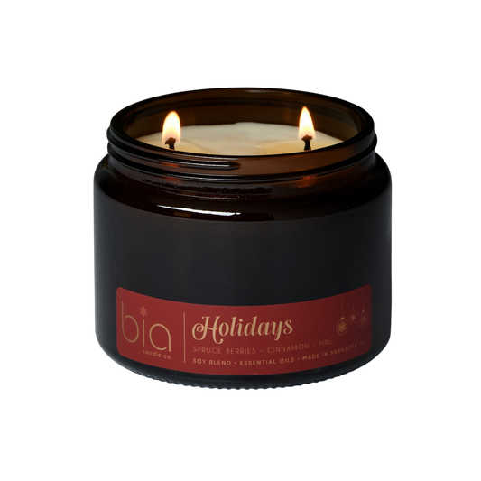Dark amber candle jar with two lit flames and “Holidays” label