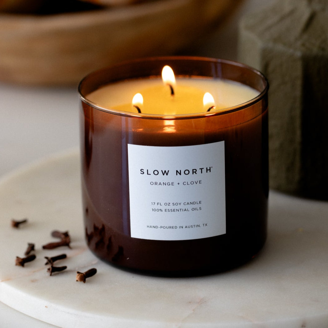 Slow North 3-wick candle in amber glass with orange-clove label and three flames