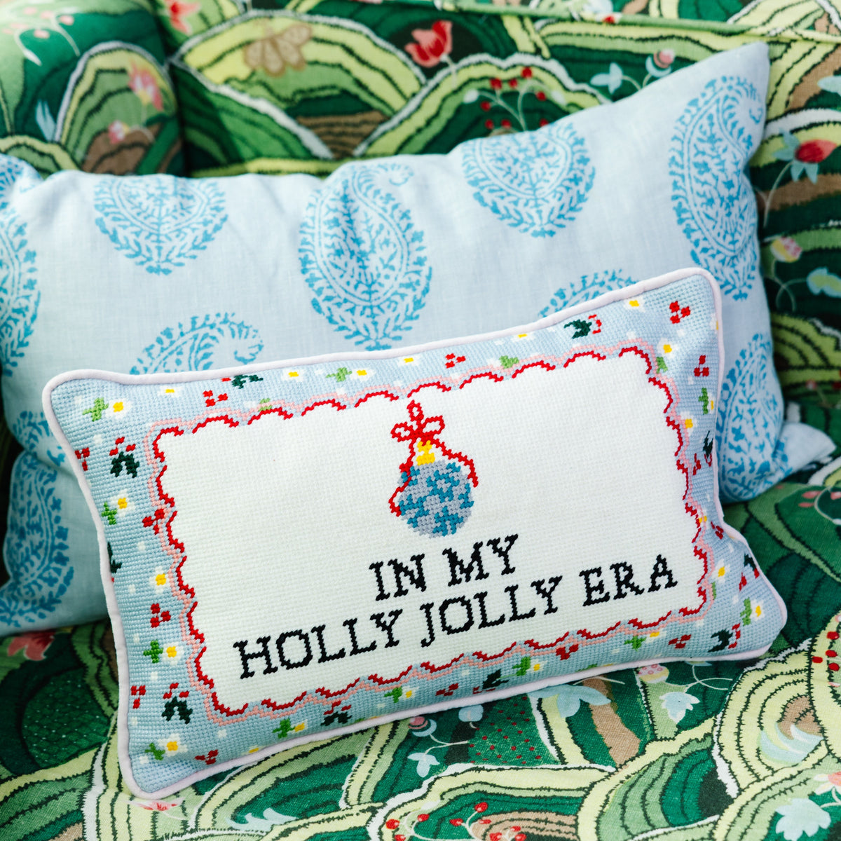 Rectangular needlepoint pillow showing 'IN MY HOLLY JOLLY ERA' in red-and-blue stitch