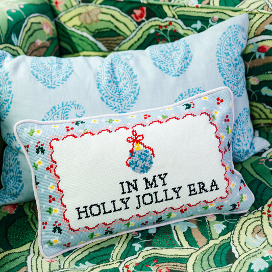 Rectangular needlepoint pillow showing 'IN MY HOLLY JOLLY ERA' in red-and-blue stitch