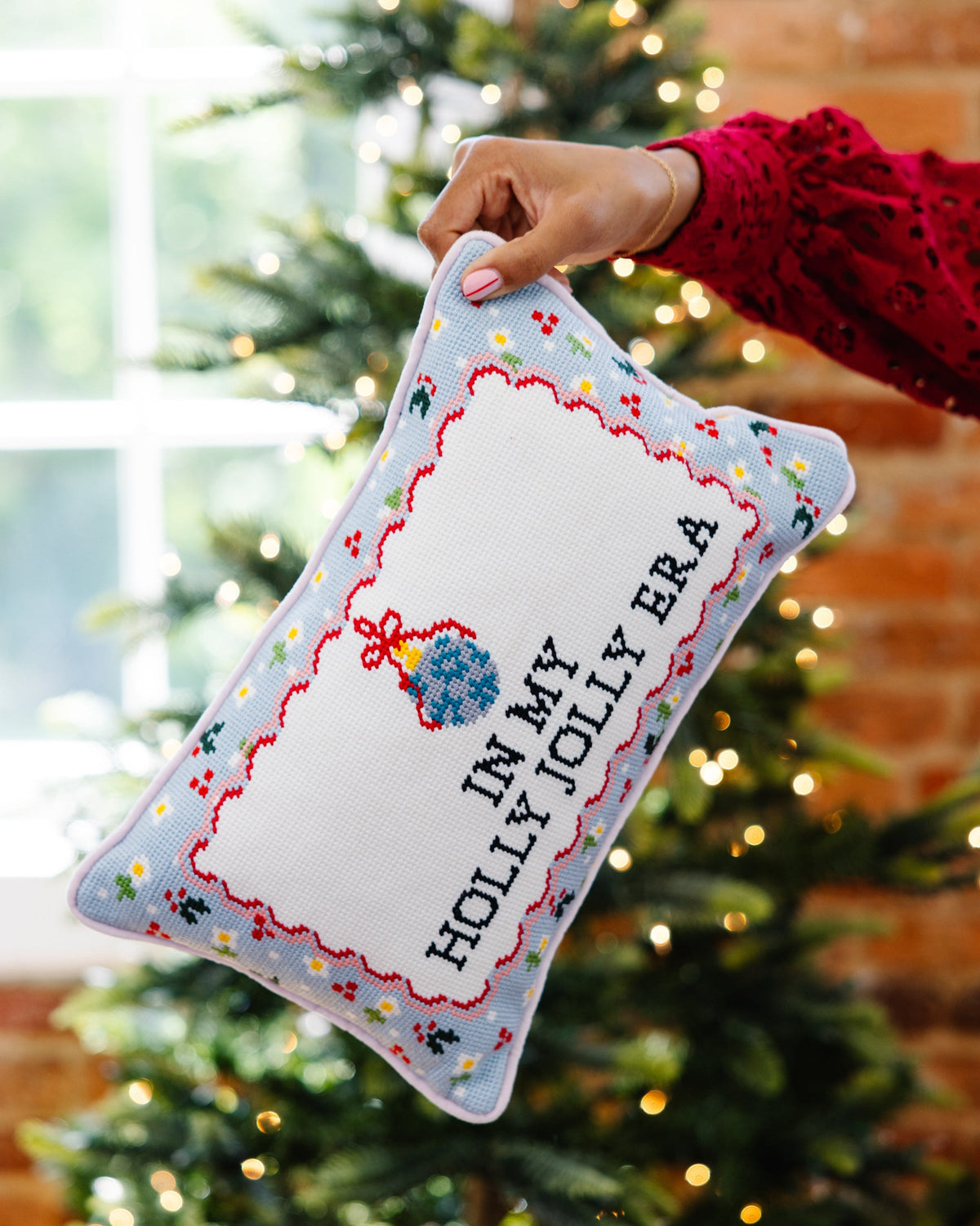 Rectangular needlepoint pillow held near a tree with 'IN MY HOLLY JOLLY ERA' text