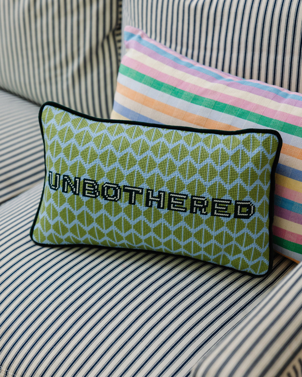 Green needlepoint throw pillow, black trim, UNBOTHERED lettering, on striped cushion