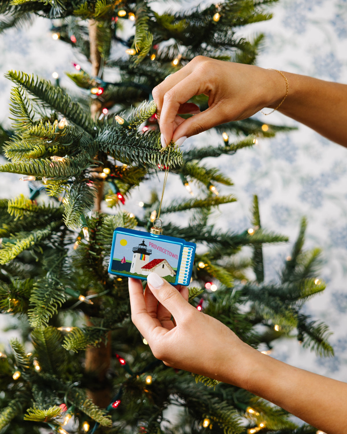 Hands placing Provincetown matchbook ornament on pine branch with gold cord