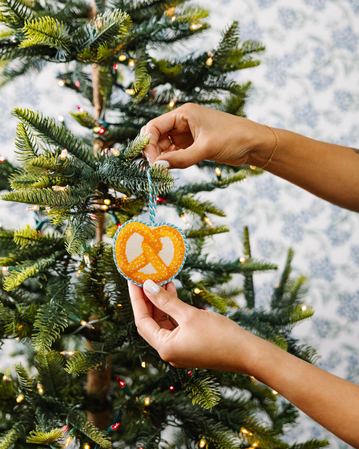 Needlepoint pretzel icon ornament being hung on a tree, orange-and-white pattern with blue loop