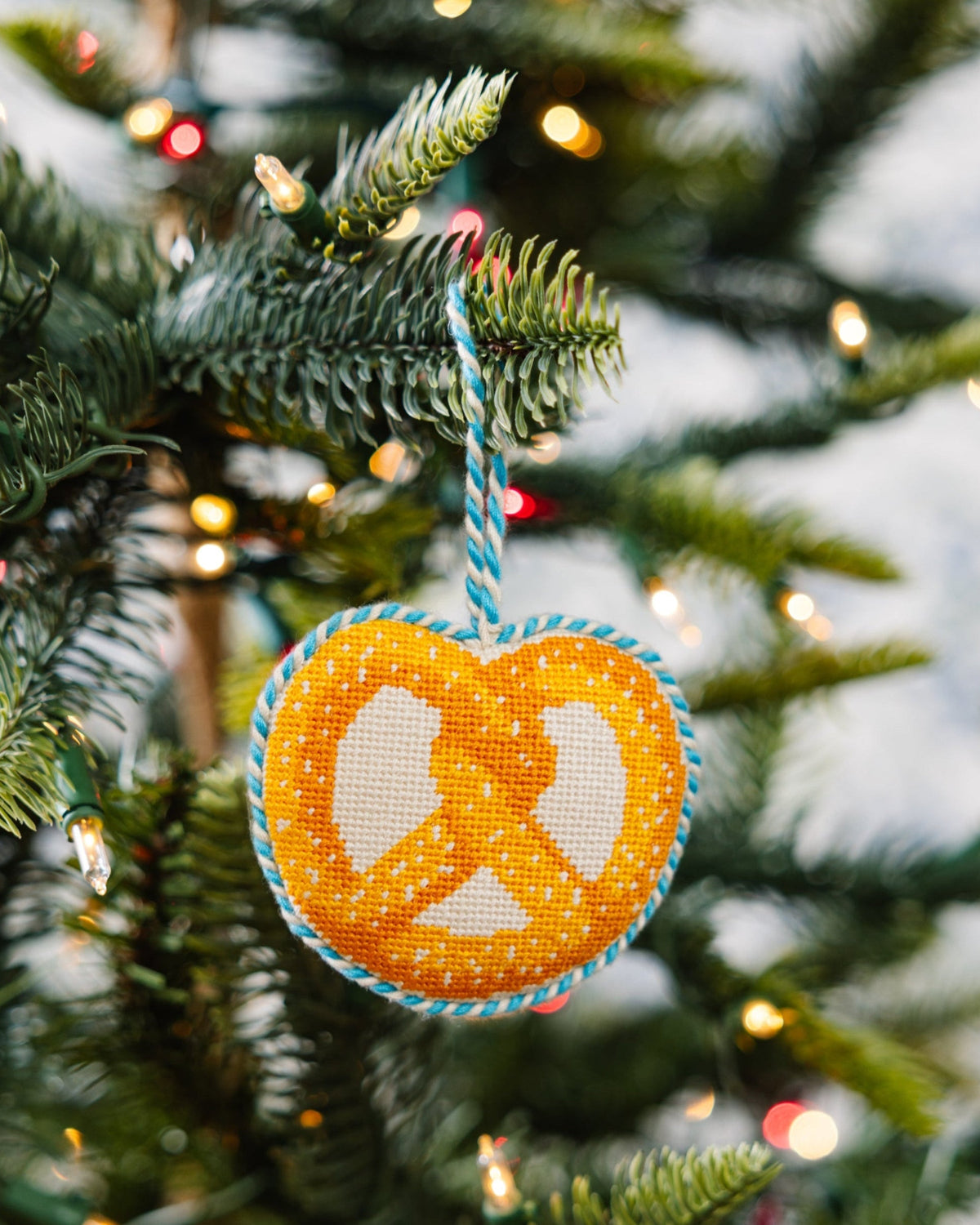 Hanging needlepoint pretzel icon ornament with orange-and-white cross-stitch and blue edging