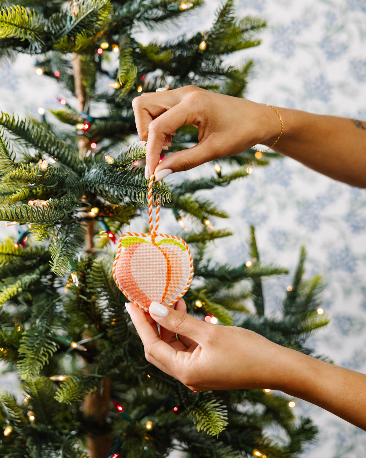 Peach needlepoint heart ornament on Christmas tree, held by hands with striped hanger