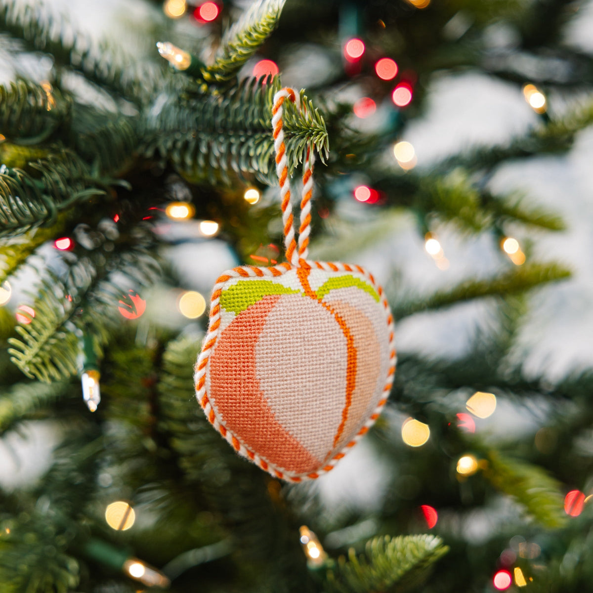 Peach needlepoint heart ornament with striped hanger hanging on a Christmas tree