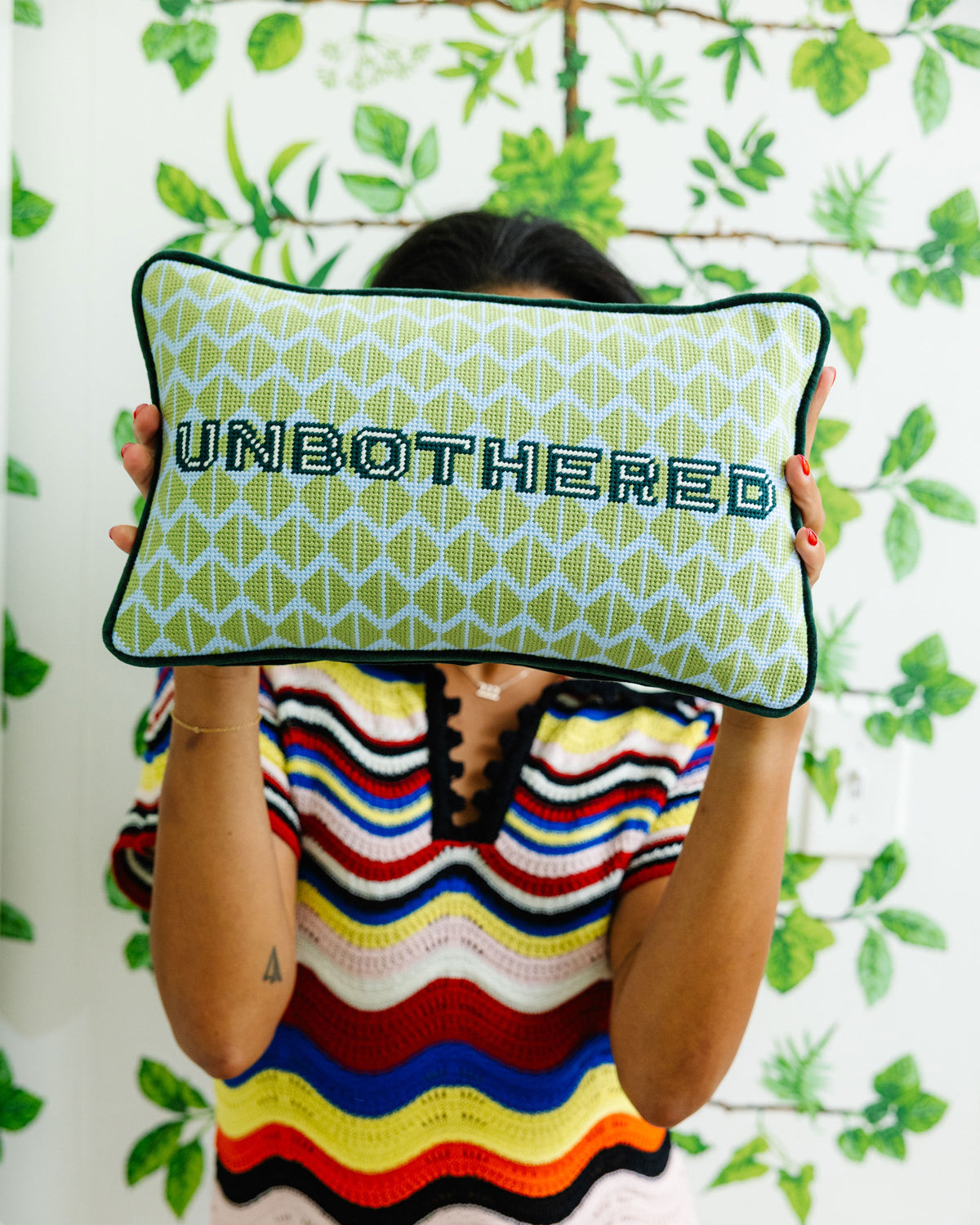 Green needlepoint pillow with black trim, UNBOTHERED lettering, held in front of plants