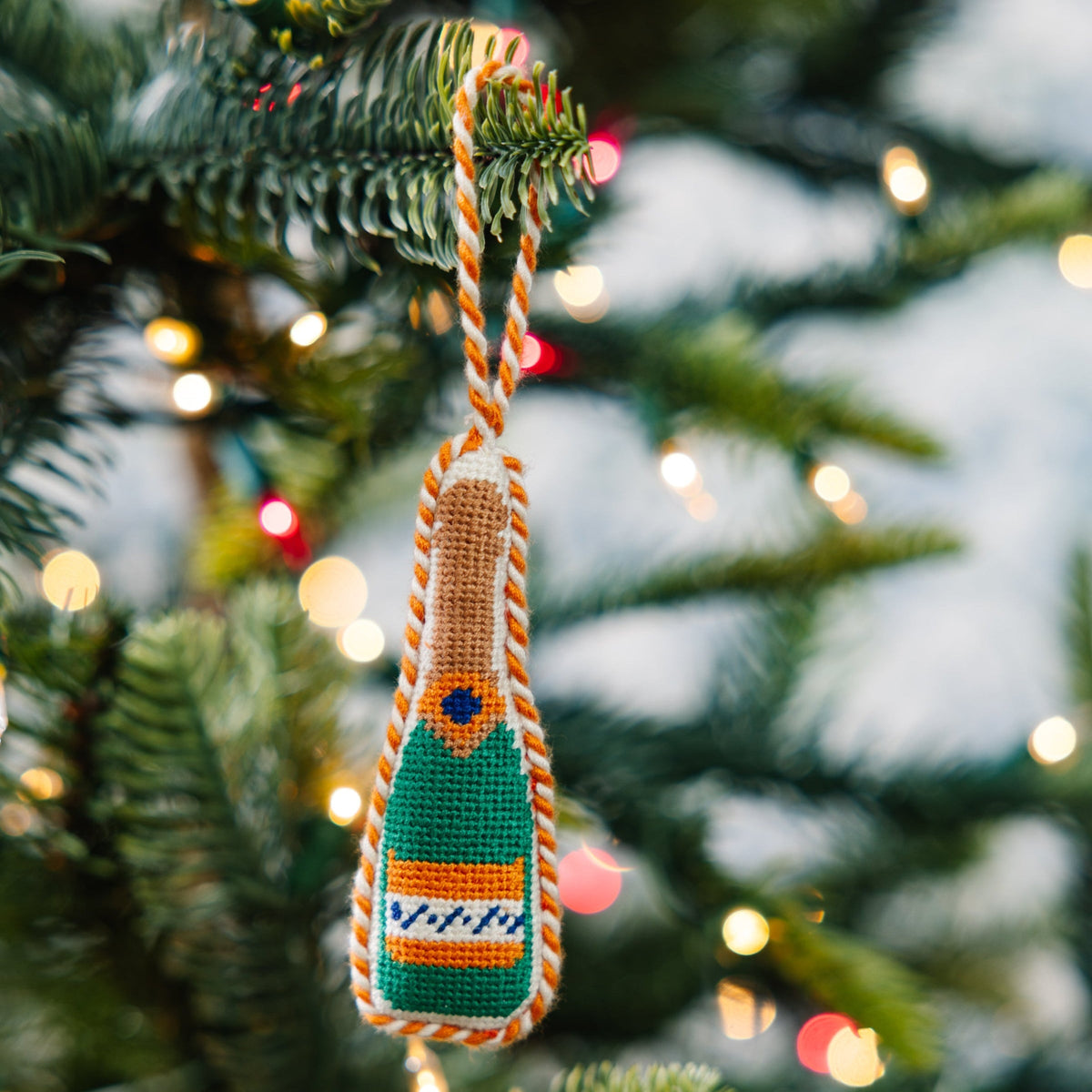 Needlepoint champagne bottle ornament with green and orange stitched label, hanging loop.