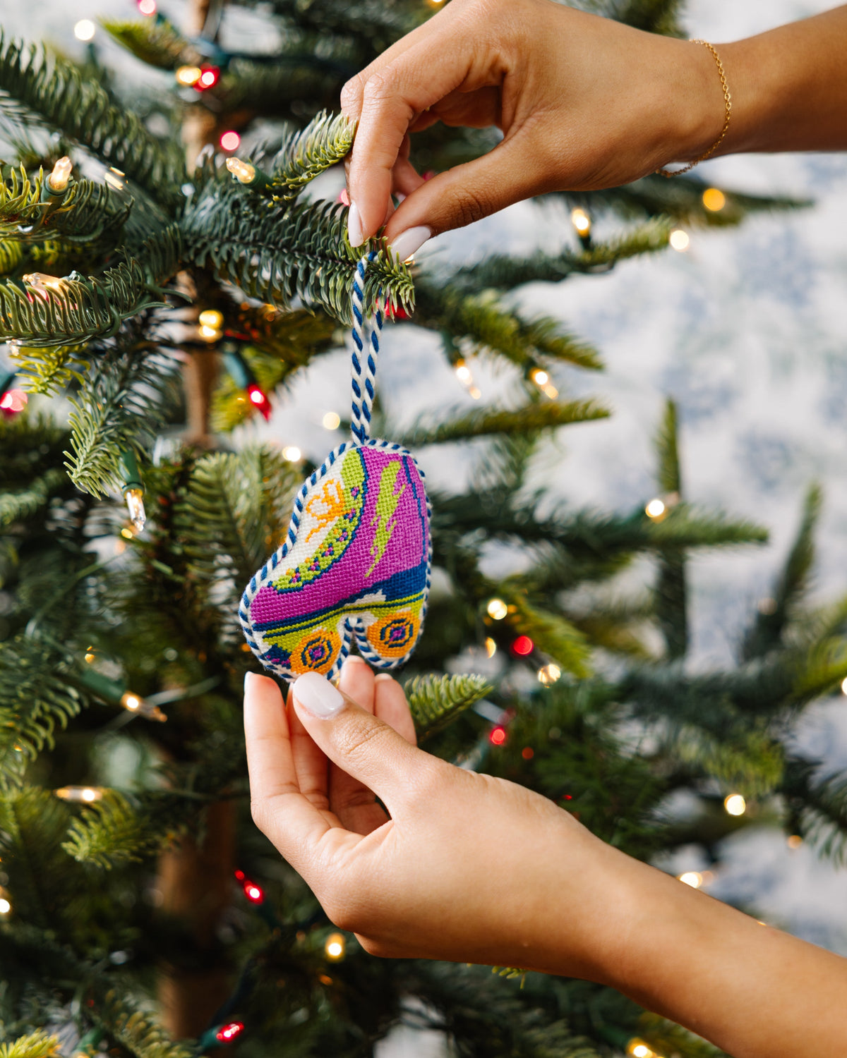 Needlepoint roller skate ornament with braided loop, hands placing it on a tree