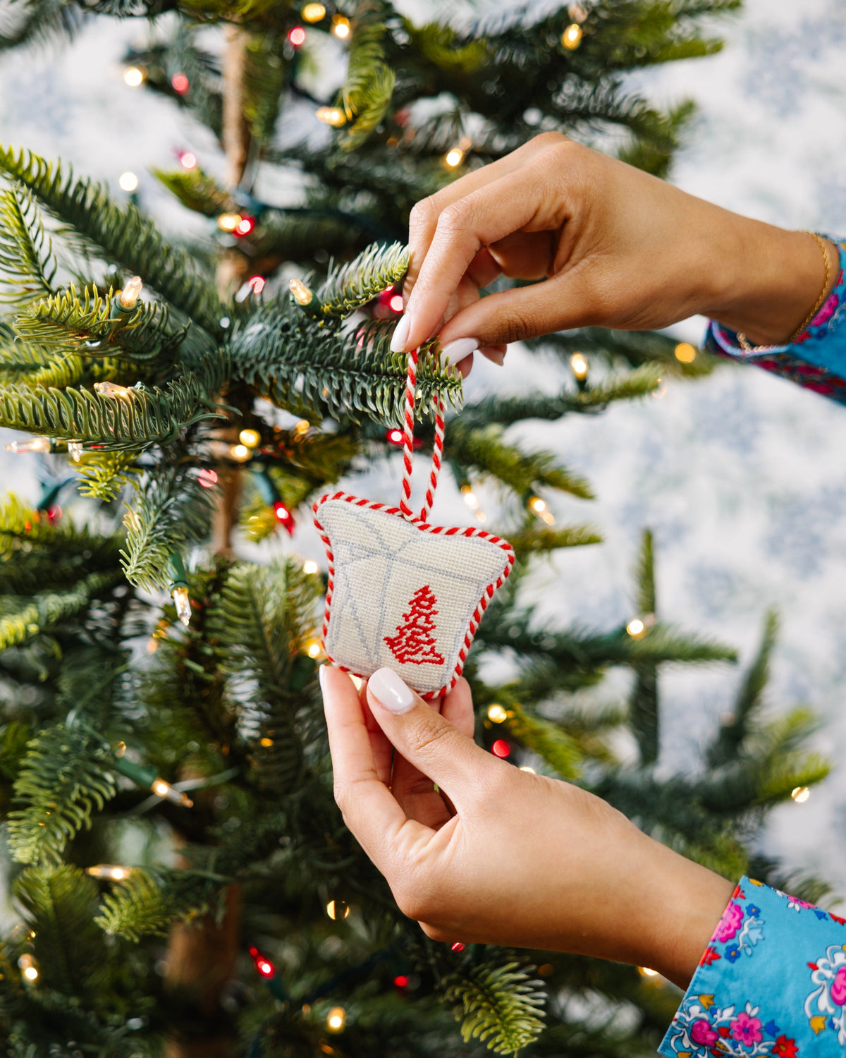Two hands hanging needlepoint takeout box ornament on a tree branch
