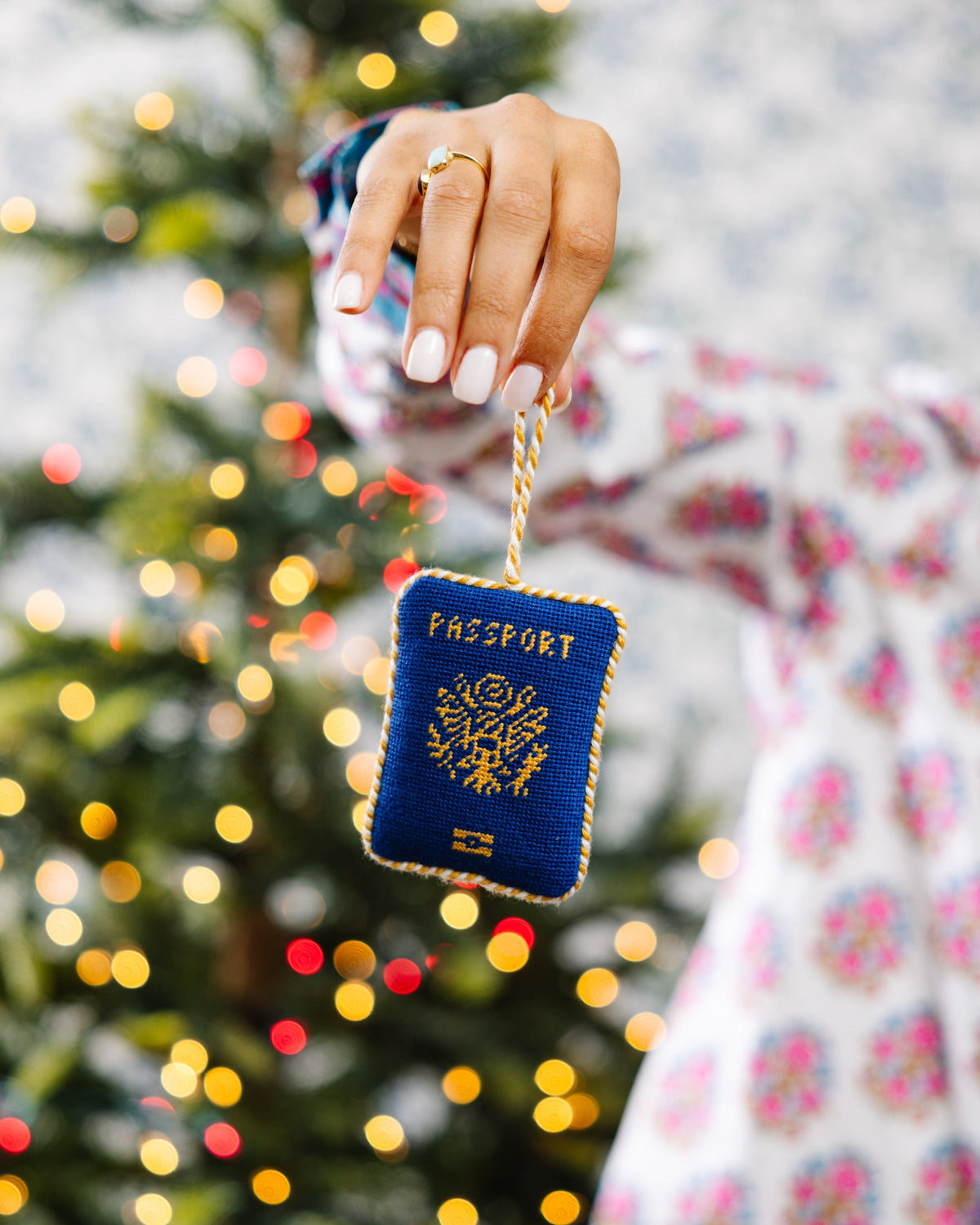 Blue needlepoint passport icon ornament held by a hand with a gold rope loop.