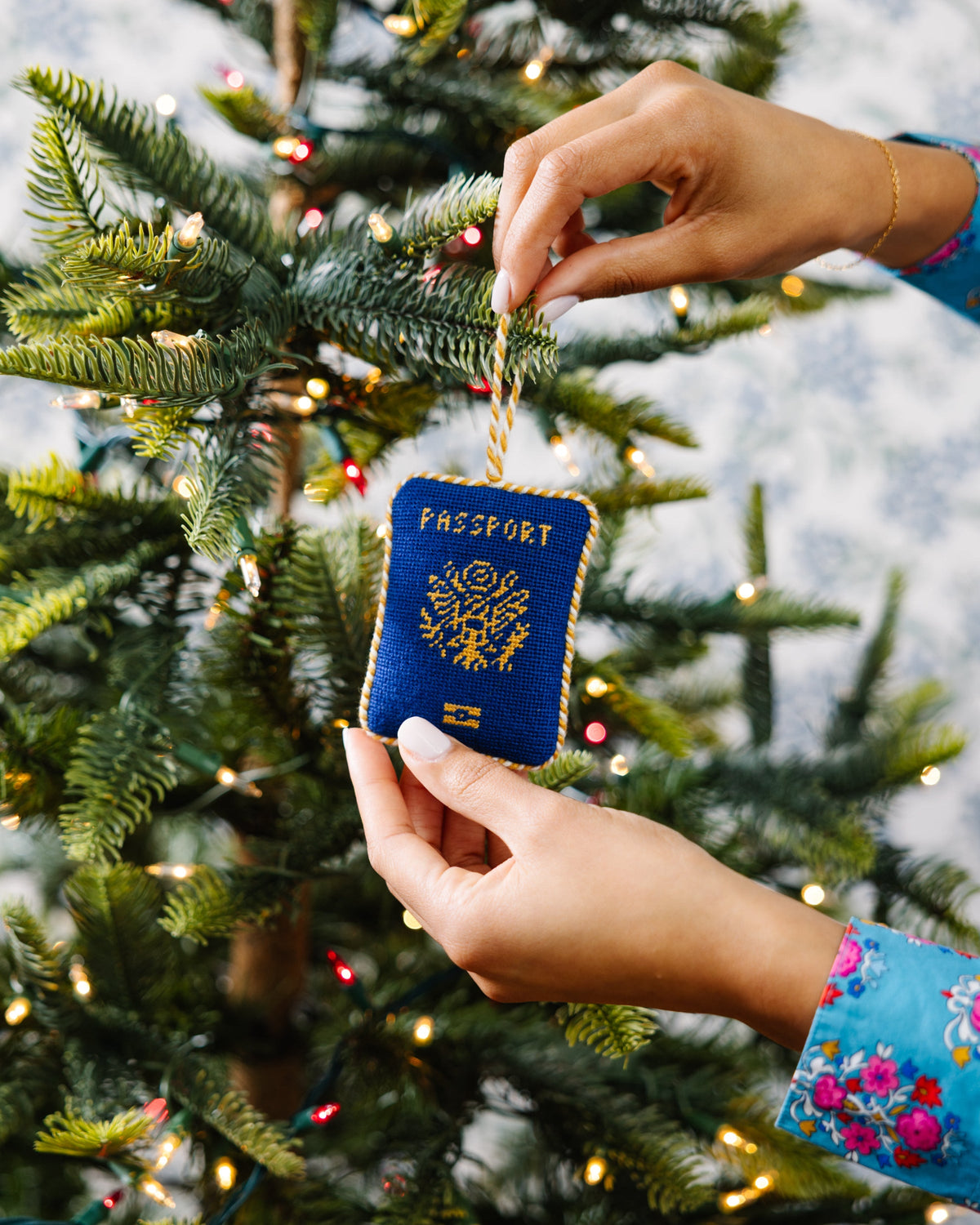 Blue needlepoint passport ornament held near fir branches, gold stitched emblem and text.