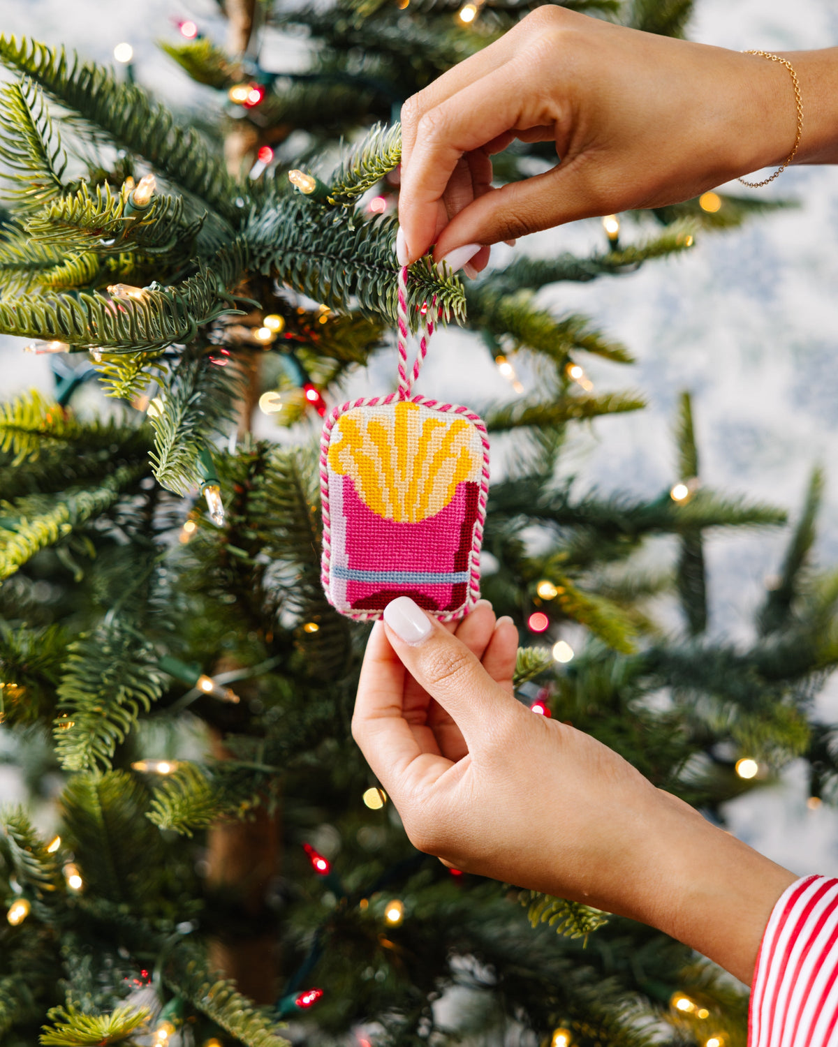 Needlepoint French fries ornament being hung on a tree branch, pink and yellow stitched square