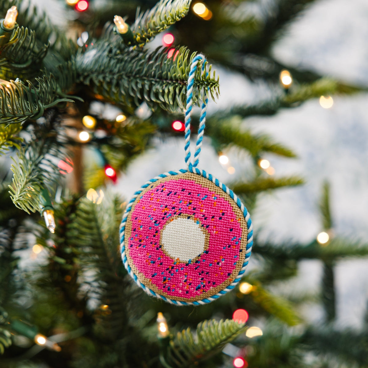 Needlepoint donut ornament in pink with sprinkles, blue trim and white center, hanging on tree
