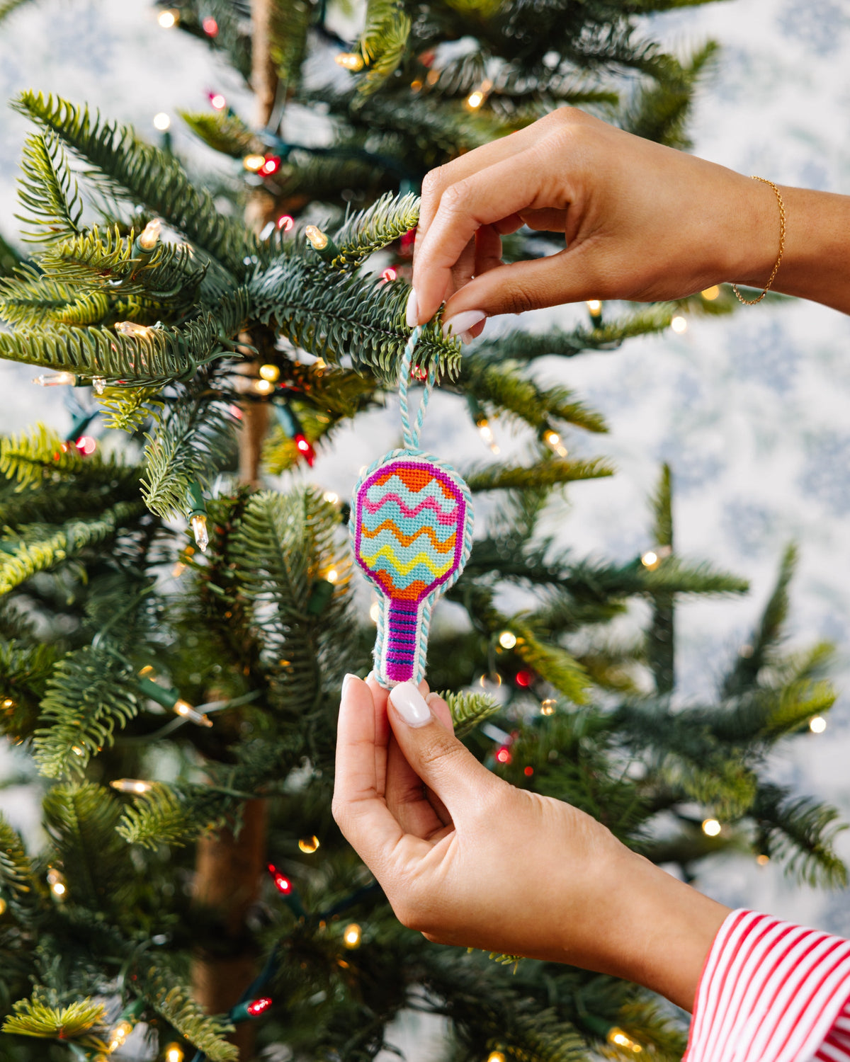 Needlepoint pickleball racket ornament being hung on a tree, multicolor zigzags.