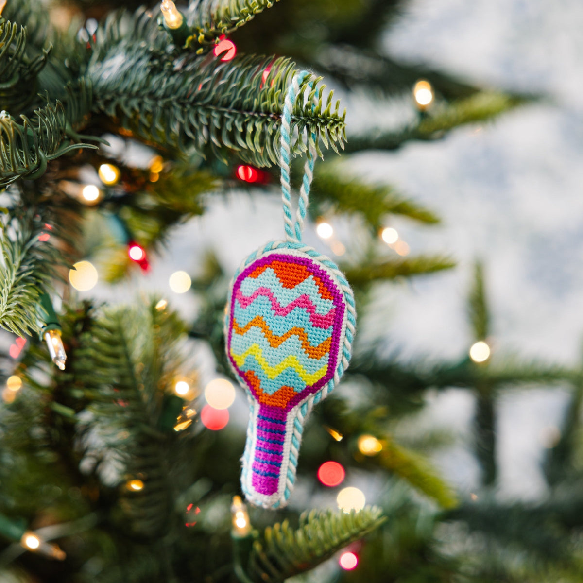 Needlepoint pickleball racket ornament hanging on a tree branch, multicolor zigzags.